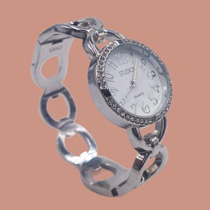 May include: A silver-toned quartz watch with a white face and crystal-studded bezel. The watch band is a series of interconnected oval links. The watch face displays the word "STUDIO" and the numbers 1 through 12.