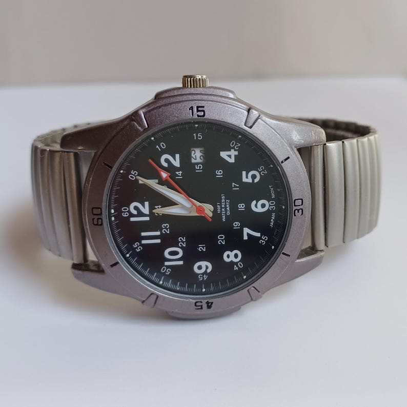 K&ouml;nnte beinhalten: Eine silberfarbene Armbanduhr mit schwarzem Zifferblatt und wei&szlig;en Ziffern. Die Uhr hat ein silbernes, dehnbares Armband und einen roten Sekundenzeiger. Das Zifferblatt zeigt das Datum und ist mit "100FT WATER RESIST QUARTZ JAPAN 30 MOVT." gekennzeichnet.