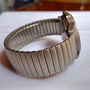 K&ouml;nnte beinhalten: Eine silberfarbene Armbanduhr mit einem dehnbaren Armband. Das Zifferblatt der Uhr ist rund und das Armband besteht aus kleinen, rechteckigen Metallgliedern. Die Uhr wird auf einer wei&szlig;en Oberfl&auml;che gezeigt.
