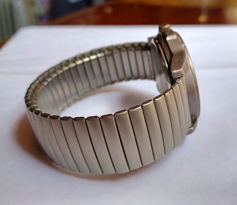 K&ouml;nnte beinhalten: Eine silberfarbene Armbanduhr mit einem dehnbaren Armband. Das Zifferblatt der Uhr ist rund und das Armband besteht aus kleinen, rechteckigen Metallgliedern. Die Uhr wird auf einer wei&szlig;en Oberfl&auml;che gezeigt.