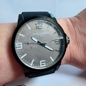 K&ouml;nnte beinhalten: Eine schwarze Armbanduhr mit rundem Geh&auml;use und dunkelgrauem Zifferblatt. Das Zifferblatt zeigt den Markennamen ROUSSEAU, wei&szlig;e arabische Ziffern bei 12, 3, 6 und 9 Uhr, wei&szlig;e Zeiger, einen d&uuml;nnen Sekundenzeiger und ein Datumsfenster bei 3 Uhr.