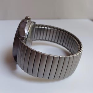 K&ouml;nnte beinhalten: Eine silberfarbene Armbanduhr mit einem ausziehbaren Metallarmband. Das Zifferblatt der Uhr ist dunkel, und das Armband besteht aus rechteckigen Gliedern. Die Uhr ist vor einem wei&szlig;en Hintergrund platziert.