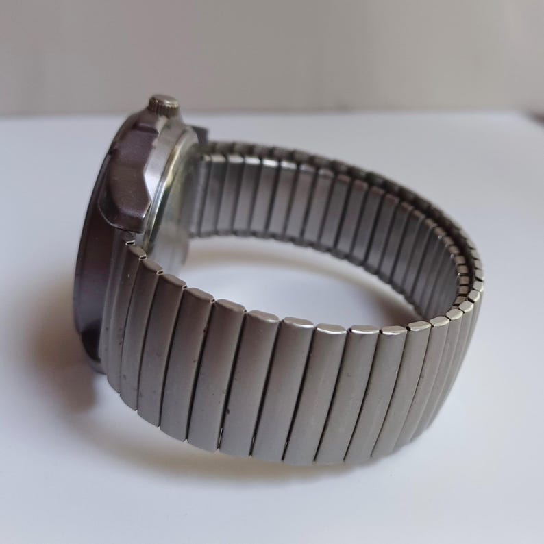 K&ouml;nnte beinhalten: Eine silberfarbene Armbanduhr mit einem ausziehbaren Metallarmband. Das Zifferblatt der Uhr ist dunkel, und das Armband besteht aus rechteckigen Gliedern. Die Uhr ist vor einem wei&szlig;en Hintergrund platziert.