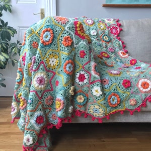 May include: A vibrant, hand-crocheted blanket with a floral design, draped over a sofa. The blanket showcases a mix of colours, including blue, orange, pink, and yellow, with a bright pink trim. This decorative blanket is a cozy addition to any home.