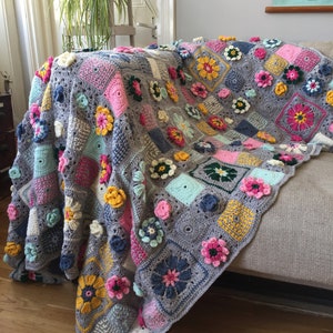May include: A handmade, grey crocheted blanket with colourful floral accents. The blanket features squares of various colours, including pink, yellow, blue, and white, each adorned with three-dimensional flower appliqués. The blanket is draped over a sofa.