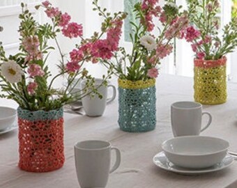 May include: A dining table set for a meal, featuring white dishes, mugs, and colorful crochet-covered vases filled with flowers. The vases are in shades of red, blue, and yellow. The table is covered with a neutral-colored tablecloth.