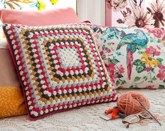 May include: A square, crocheted pillow with a granny square design in red, white, gray, and yellow. A ball of yarn, crochet hook, and other craft supplies sit nearby. Other pillows with floral patterns are in the background.