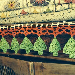 May include: A stack of handmade crochet blankets in various colors, including cream, gray, and brown, with floral and geometric patterns. A decorative orange and green crochet garland with tree shapes hangs in front.