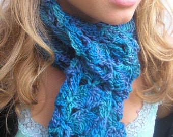 May include: A hand-knitted scarf in shades of blue and turquoise. The scarf features a textured, openwork design, with a scalloped edge. The yarn appears to be a soft, bulky weight. The scarf is draped around the neck.