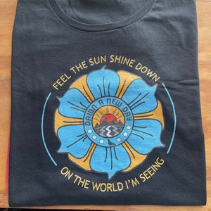 May include: Black t-shirt with a blue and gold flower design. The flower has a circle in the center with the text "Dawn a New Day" and a sunrise graphic. The text "Feel the sun shine down on the world I'm seeing" surrounds the flower.