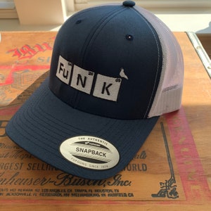 May include: Navy blue Snapback hat with a white mesh back. The hat features the word "FUNK" in a periodic table style, with the brand "YUPOONG" on the front. The hat is sitting on a wooden surface.