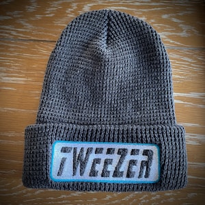 May include: A gray knit beanie with a folded brim. The beanie features a rectangular patch with the word "WEEZER" in white block letters, outlined in light blue. The hat is made of a textured knit material.