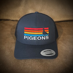 Könnte beinhalten: Eine dunkelblaue Trucker-Kappe mit schwarzem Netzrücken. Die Vorderseite der Kappe zeigt ein weiß gesticktes Design eines Taubens mit einem Streifenmuster in Sonnenuntergangsfarben und dem Wort "PIGEONS". Die Kappe hat ein schwarzes Etikett auf der Rückseite, auf dem "The Authentic Snapback Yupoong Since 1974" steht.