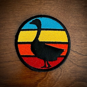 May include: A black silhouette of a goose in front of a sunset-colored background. The background is a circle with stripes of blue, yellow, orange, and red.
