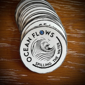May include: A white round sticker with black text that reads "Ocean Flows The Water Spilling" with a blue wave graphic and a blue drop of water.