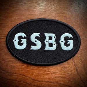 May include: Black oval patch with the embroidered letters "GSBG" in light blue. The patch has a black border and is set against a wooden background. The letters have a unique, stylized design.