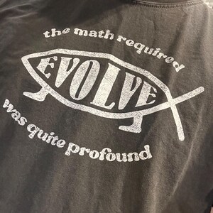 May include: Dark gray t-shirt with a white graphic. The graphic features a fish shape with the word "EVOLVE" inside. Above the fish is the text "the math required" and below it reads "was quite profound."
