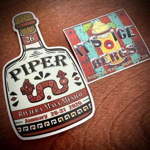 May include: Two stickers on a wooden surface. One is shaped like a bottle with the word "PIPER" and "Riviera Maya Mexico January 28-31 2026" printed on it. The other sticker says "IT'S ICE BERGS Riviera Maya Mexico January 28-31 2026".