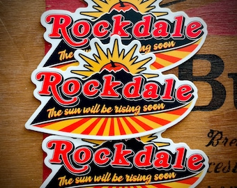 May include: Three stickers with a sunburst design and the text "Rockdale" in red letters. The text "The sun will be rising soon" is also included on the bottom two stickers.