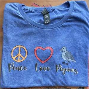 Könnte beinhalten: Ein blaues T-Shirt mit einem Peace-Zeichen, einem Herzen und einer Taube. Der Text "Peace Love Pigeons" ist auf dem T-Shirt aufgedruckt.