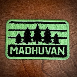 Puede incluir: Un parche bordado rectangular con fondo verde claro y rayas horizontales negras. El diseño muestra cinco siluetas de pinos negros de diferentes tamaños. La palabra MADHUVAN está bordada en letras mayúsculas de color verde claro.