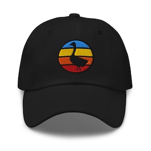 May include: A black baseball cap featuring an embroidered circular patch on the front. The patch displays a black silhouette of a goose centered against a horizontal striped background of blue, yellow, orange, and red colors, mimicking a sunset.