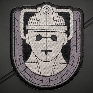 May include: A gray and black embroidered patch featuring a stylized image of a Dalek, a fictional robot from the British science fiction television series Doctor Who.