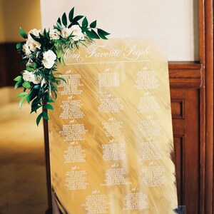 Op de afbeelding: Een gouden acryl bord met witte kalligrafie met een lijst van namen voor een huwelijksreceptie. Het bord is versierd met een bloemstuk van witte rozen en groene bladeren.
