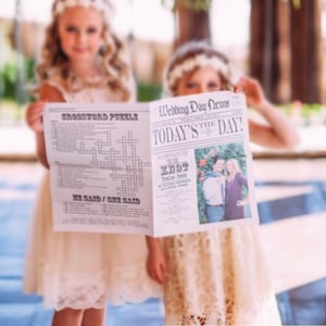 May include: Two girls in white dresses hold a newspaper-style wedding announcement with a crossword puzzle on one side and a wedding photo on the other. The headline reads "Wedding Day News" and "Today's the Day!"