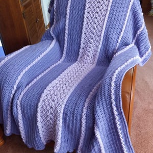 May include: A handmade crochet blanket in shades of periwinkle and lavender, draped over a chair. The blanket features vertical stripes with textured patterns and a scalloped edge. The color palette is soft and inviting, perfect for home decor.