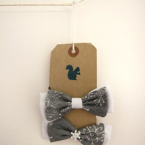 May include: Two gray and white bow ties with snowflake and geometric patterns hanging from a brown tag with a squirrel silhouette. The tag is attached to a string.