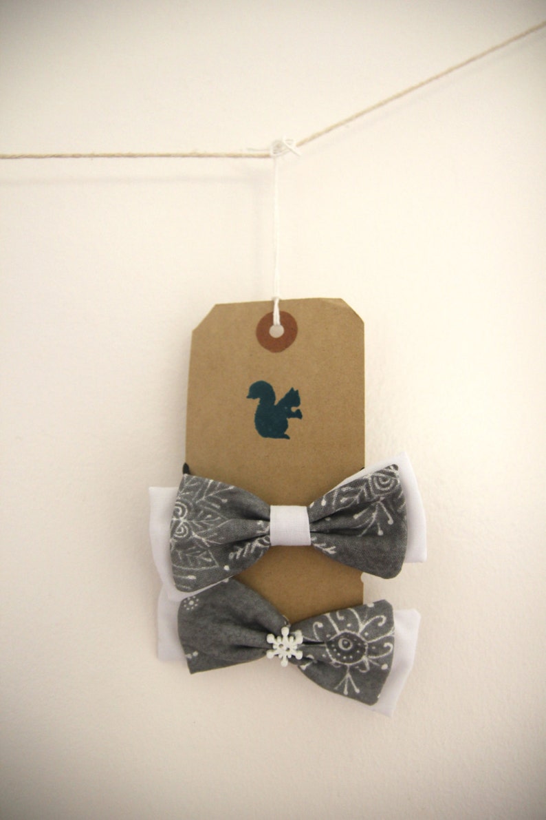 May include: Two gray and white bow ties with snowflake and geometric patterns hanging from a brown tag with a squirrel silhouette. The tag is attached to a string.