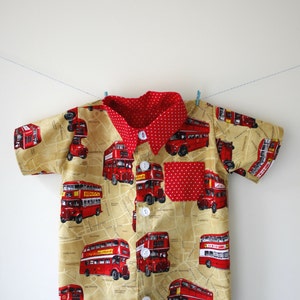 May include: A short-sleeved button-down shirt for children with a red and white polka dot collar and a pocket. The shirt is made of a beige fabric with a pattern of red double-decker buses.