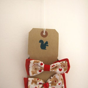 May include: Two red and white bow ties with a reindeer pattern. The bow ties are hanging from a brown tag with a squirrel silhouette.