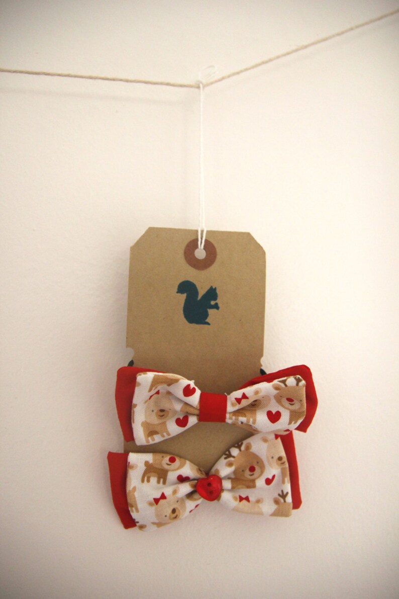 May include: Two red and white bow ties with a reindeer pattern. The bow ties are hanging from a brown tag with a squirrel silhouette.
