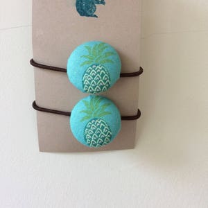 Könnte beinhalten: Zwei braune Haargummis mit türkisfarbenen Stoffknöpfen mit Ananas-Motiv.