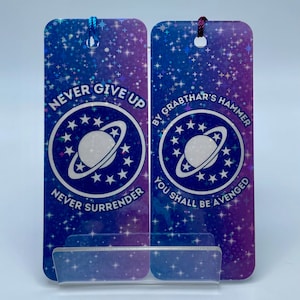 May include: Two galaxy-themed bookmarks with a blue and purple background. The left bookmark says "Never Give Up" and "Never Surrender" with a planet and stars. The right bookmark says "By Grabthar's Hammer" and "You Shall Be Avenged" with a planet and stars.