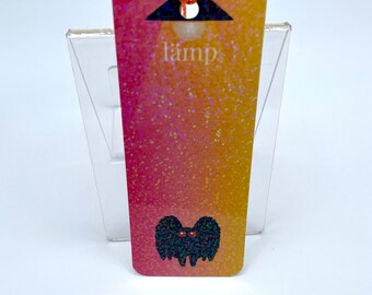 May include: A pink and yellow gradient bookmark with a black silhouette of a monster with red eyes. The bookmark has a hole at the top and the word "lamp" printed on it.