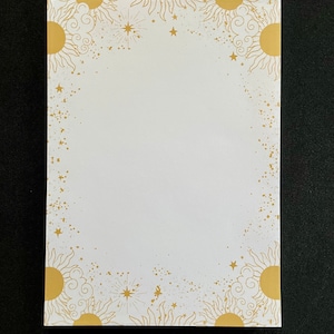 May include: A white notepad with a gold border featuring a repeating pattern of suns and stars. The suns have a stylised design with rays and clouds.