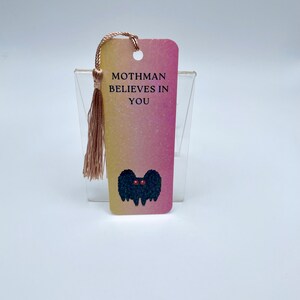 May include: A pink and yellow glitter bookmark with a black mothman illustration and the text "Mothman Believes in You".
