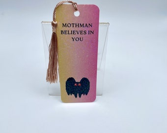 May include: A pink and yellow glitter bookmark with a black mothman illustration and the text "Mothman Believes in You".