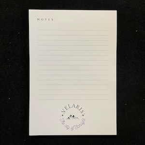 May include: A white notepad with lined paper and the words "NOTES" at the top. The bottom of the notepad has a black and white logo that says "VELARIS The city of Starlight".