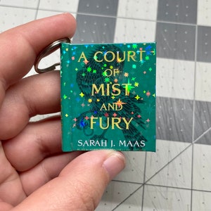 Puede incluir: Un llavero de libro verde con acabado holográfico. La portada del libro presenta una ilustración en blanco y negro de un pájaro con el título "A Court of Mist and Fury" y el nombre del autor "Sarah J. Maas".