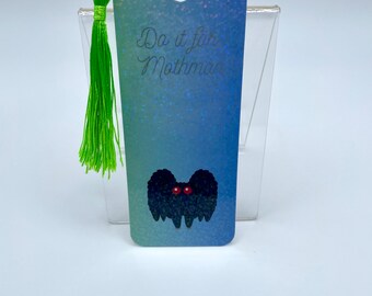 May include: A bookmark with a gradient blue and green background and the text "Do it for Mothman." It features a black, furry Mothman illustration with red eyes. A green tassel hangs from the top.