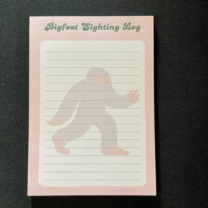 May include: Pink notepad with a green title that says "Bigfoot Sighting Log". A silhouette of Bigfoot is printed on the notepad.