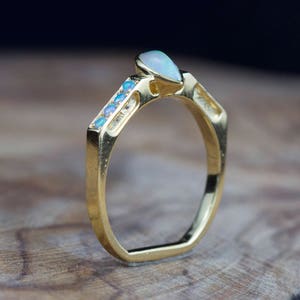 May include: A gold ring with a pear-shaped opal gemstone and blue accent stones. The ring has a geometric design and is set on a wooden surface.