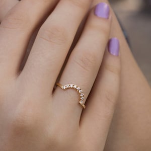 Könnte beinhalten: Ein goldener Ring mit einer geschwungenen Linie aus kleinen, klaren Steinen. Der Ring wird an einem Finger mit helllila Nagellack getragen.