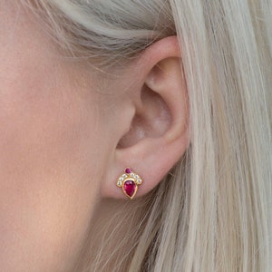 Pode incluir: Um par de brincos de ouro com uma gema de rubi em forma de l&aacute;grima e um halo de gemas brancas menores.