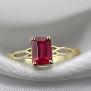 May include: A gold ring featuring a red emerald-cut gemstone set in a delicate, interwoven band.