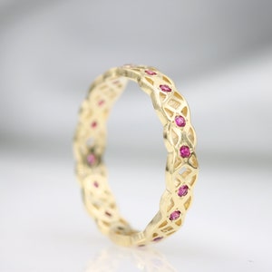 May include: A gold band ring with a delicate, intricate design featuring a woven pattern. The ring is accented with small, pink gemstones.